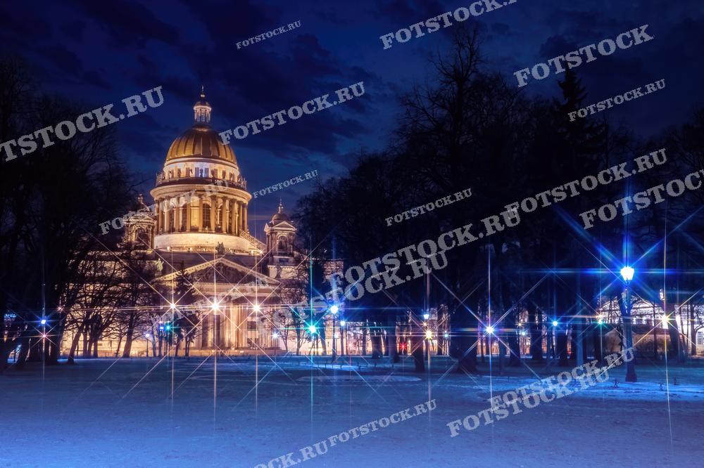 На фотографии изображен Исаакиевский собор в Санкт-Петербурге в ночное время. Подсветка подчеркивает величественные формы здания, создавая эффектный контраст с темным небом. Зимний пейзаж вокруг собора дополняет атмосферу. Исаакиевский собор является одним из самых известных архитектурных памятников города.